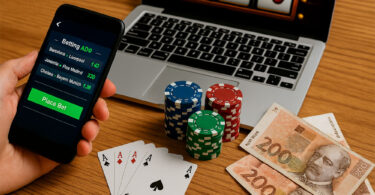 Ruka drži mobitel s aplikacijom za klađenje, ispred laptopa s automatima, žetonima, kartama i novčanicama od 200 kuna na drvenom stolu.